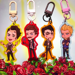May include: Four colorful keychains featuring cartoon characters with different hairstyles and clothing. The keychains are attached to silver, black, and pink key rings. The keychains are displayed against a background of red roses.