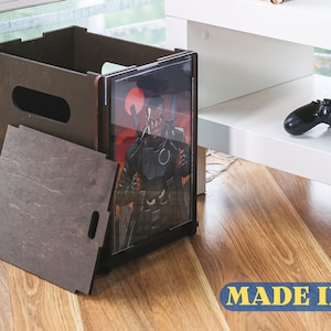 Puede incluir: Una caja de almacenamiento de madera con una tapa extraíble y un panel frontal de plástico transparente. La caja está sentada en un suelo de madera y tiene un cómic dentro. El texto "MADE IN UA" es visible en el suelo.