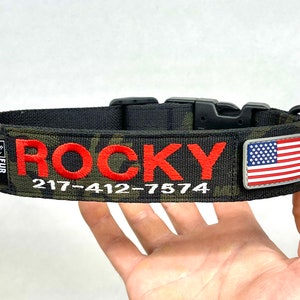 May include: A black and green camouflage dog collar with a red embroidered name "ROCKY" and phone number "217-412-7574". The collar has a black buckle and a small American flag patch.