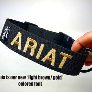 May include: Black dog collar with the name "ARIAT" embroidered in a light brown/gold color. The collar has a black plastic buckle and a metal ring. The brand "FUR FORGE" is printed on a small label.