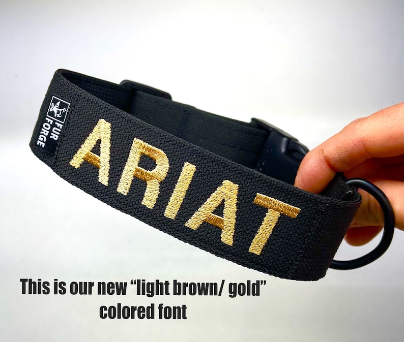 May include: Black dog collar with the name "ARIAT" embroidered in a light brown/gold color. The collar has a black plastic buckle and a metal ring. The brand "FUR FORGE" is printed on a small label.