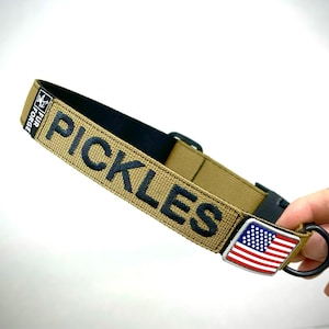 May include: A tan dog collar with black stitching and the word "PICKLES" embroidered in black. The collar has a black buckle and a silver metal tag with an American flag design.