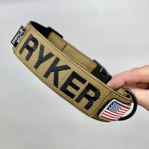 May include: Tan dog collar with black stitching and the word "RYKER" embroidered in black. The collar has a black buckle and a small American flag charm.