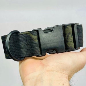 May include: A black dog collar with a camouflage pattern in shades of green and brown. The collar features a black plastic buckle and a black metal D-ring for leash attachment. The collar is held in a hand.