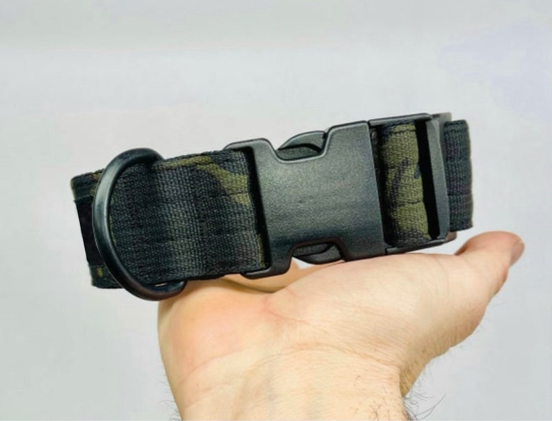 May include: A black dog collar with a camouflage pattern in shades of green and brown. The collar features a black plastic buckle and a black metal D-ring for leash attachment. The collar is held in a hand.