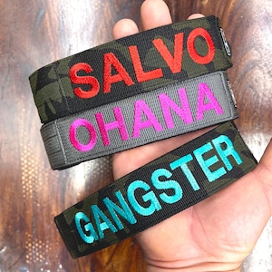 May include: Three embroidered wristbands with text. The top band is camouflage with the word "SALVO" in red. The middle band is gray with "OHANA" in pink. The bottom band is camouflage with "GANGSTER" in teal.