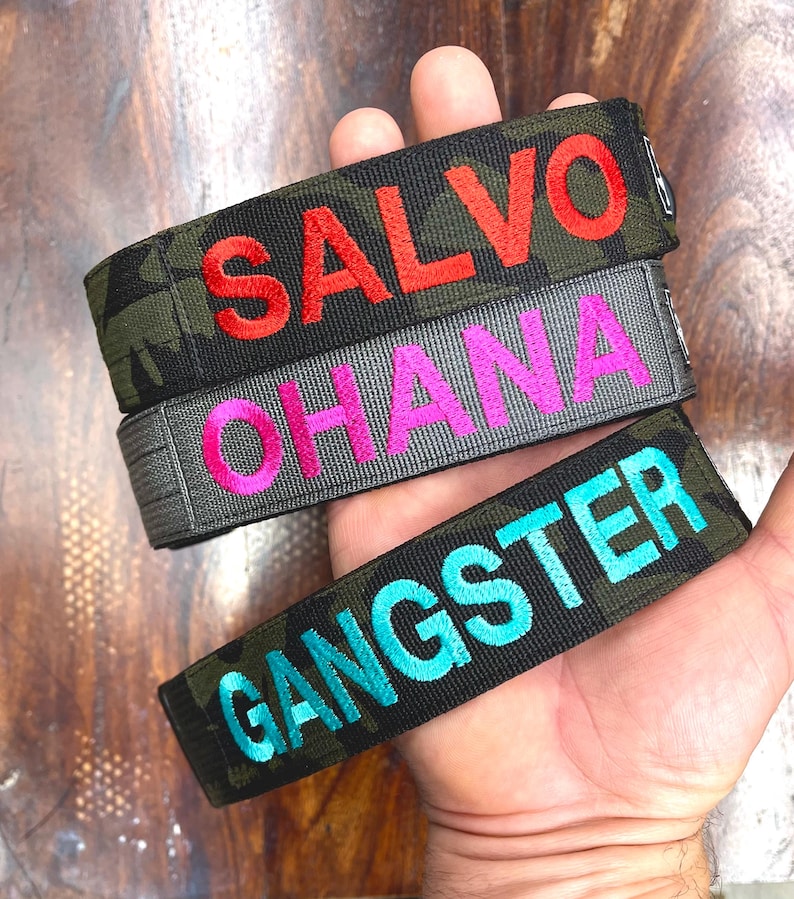 May include: Three embroidered wristbands with text. The top band is camouflage with the word "SALVO" in red. The middle band is gray with "OHANA" in pink. The bottom band is camouflage with "GANGSTER" in teal.