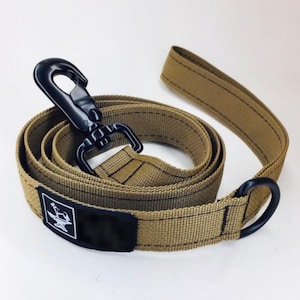 May include: A tan dog leash with a black metal clip and a black metal ring. The leash has a black patch with a white logo of an anvil and a hammer.