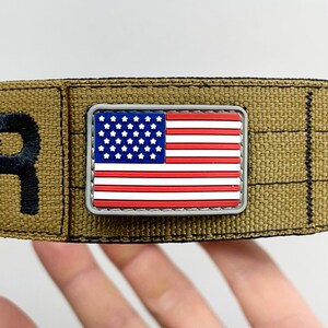 May include: A small, silver-colored patch with a red, white, and blue American flag design. The patch is attached to a brown fabric strap.