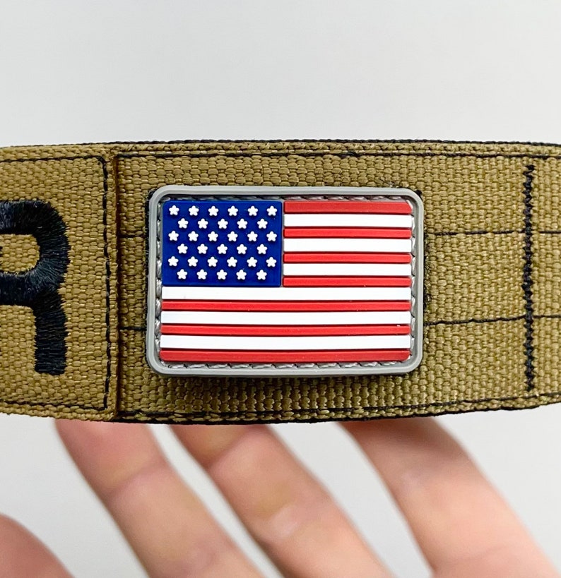 May include: A small, silver-colored patch with a red, white, and blue American flag design. The patch is attached to a brown fabric strap.
