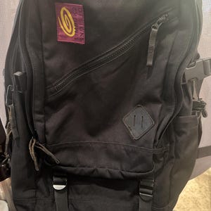 May include: A black backpack featuring a rectangular maroon patch with the yellow Timbuk2 Designs logo. The bag includes a diagonal zippered front pocket, a diamond-shaped lash tab, and two adjustable black straps with buckles at the base.