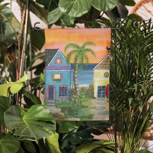 May include: A colorful garden flag with a sunset sky, two houses, a palm tree, and a beach scene. The houses are painted in shades of blue, yellow, and purple. The palm tree has green leaves and a brown trunk. The beach is a light blue color with white waves.