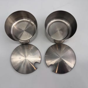 May include: Two circular stainless steel containers with matching lids. Each lid features a small semi-circular cutout along the edge, designed to accommodate a spoon. The metal has a brushed finish with a visible circular grain pattern on the lids.