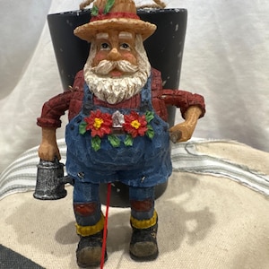 May include: A hanging ornament depicting a bearded figure wearing a straw hat, a red plaid shirt, and blue overalls with red poinsettia floral accents. The figure holds a small silver watering can in one hand and has a red string hanging below.