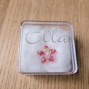 Pode incluir: Uma caixa de plástico transparente com um interior de algodão branco. A caixa contém uma estrela rosa com flocos dourados e o nome "Ella" está escrito na caixa em letras douradas.