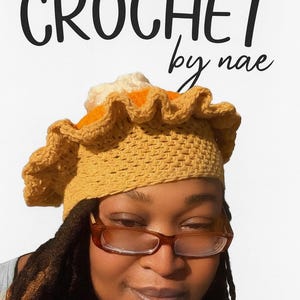 May include: A handmade crochet hat in a pie crust design with a yellow-brown color. The hat has a ruffled edge and a white and orange filling. The text "CROCHET by nae" is displayed above the hat.
