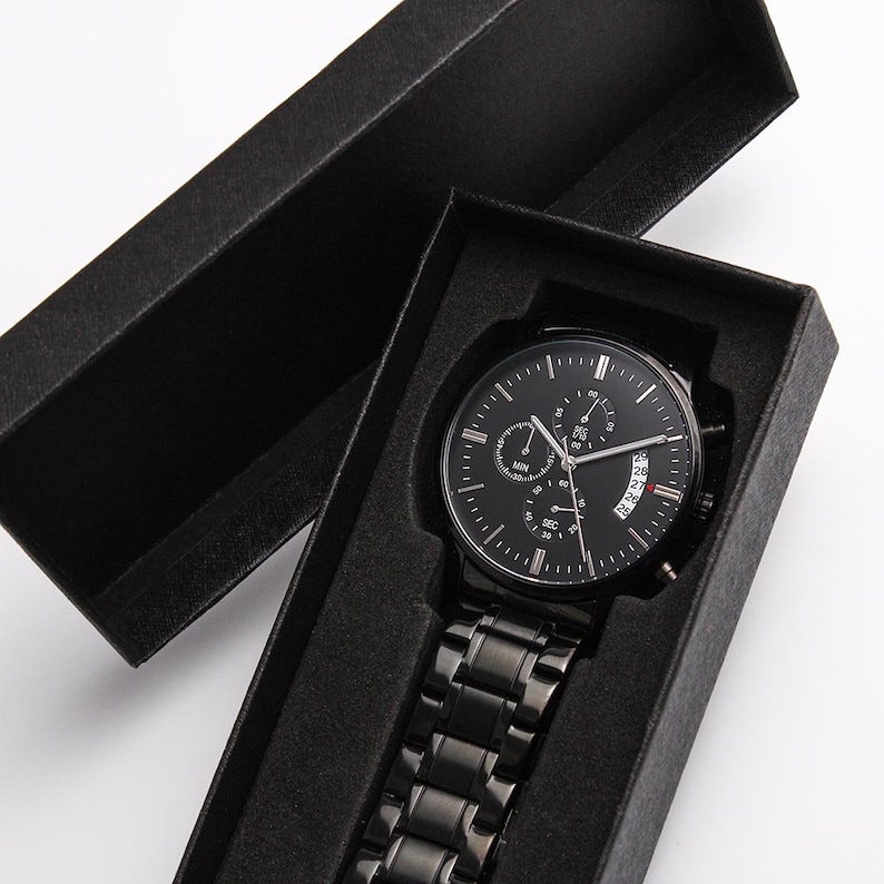 May include: A black watch with a black metal band in a black box. The watch face has a black background with silver hands and markers. The watch has a date window and a chronograph function.