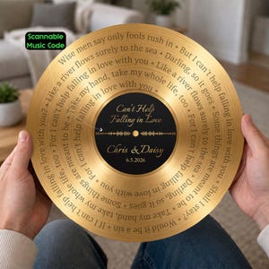 Puede incluir: Un disco de vinilo decorativo circular de color dorado con la letra de Can't Help Falling in Love grabada en espiral. La etiqueta central negra muestra el título de la canción, los nombres Chris & Daisy, la fecha 6.5.2026 y un código musical escaneable.