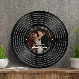 Op de afbeelding: Een zwarte vinylplaat met een wit ontwerp en een foto van een stel in het midden. De plaat heeft de tekst "Can't Help Falling in Love" en de namen van het stel en de trouwdatum.