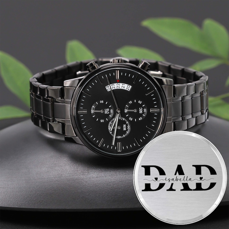 May include: A black chronograph watch with a black leather band and a silver-toned metal case. The watch face has a black background with white markings and a date window. The watch is accompanied by a silver-toned metal disc with the word "DAD" engraved on it, along with the name "isabella" and two hearts.