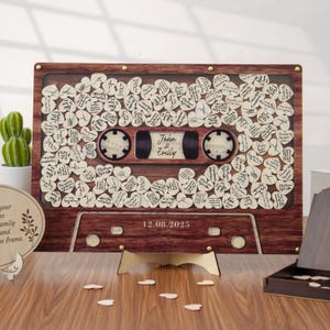 May include: A wooden cassette tape guest book with a clear front, filled with heart-shaped wooden pieces for guests to sign. The tape reel area has the names "John & Emily" and the date "12.08.2025".