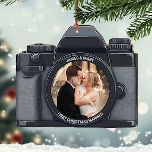 Puede incluir: Un adorno de cámara negro y plateado con una foto de una pareja besándose dentro del objetivo. El adorno dice "James & Mary" y "Primera Navidad Casados".
