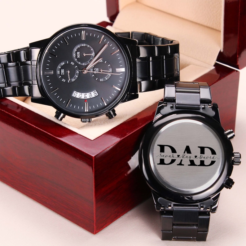 May include: Two black wristwatches in a red wooden box. The watch on the left has a black face with silver hands and markers. The watch on the right has a silver face with the word "DAD" engraved on it. The text "Sarah Kay David" is engraved below the word "DAD".