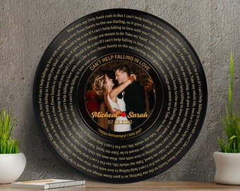 Op de afbeelding: Een zwarte vinylplaat met gouden letters die "Can't Help Falling in Love" lezen met een foto van een stel in het midden. De foto is omringd door een gouden hart met de namen "Micheal" en "Sarah" en de datum "07.03.2023". De tekst "Happy Anniversary! I love you!" staat onder de foto.