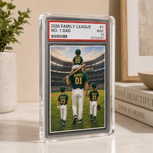 May include: A rectangular, clear acrylic display case containing a sports-style trading card. The card features an illustration of a person carrying a child on their shoulders, flanked by two other children on a baseball field, with the text 2026 FAMILY LEAGUE NO. 1 DAD.