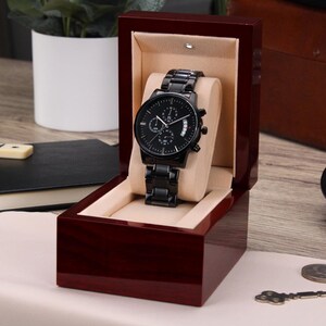 May include: A black chronograph wristwatch with a black leather band is displayed in a dark wood box with a cream-colored interior.