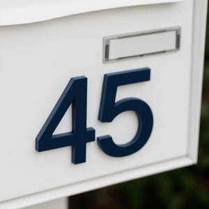 以下が含まれることがあります： 長方形のスロットと小さな鍵穴が付いた白い郵便受け。数字の「45」は濃い青色で表示され、数字の上に小さな長方形のスペースがあります。郵便受けは白い柱に取り付けられています。