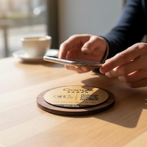 Op de afbeelding: Een ronde houten onderzetter met een goudkleurig middenstuk met het Google-logo, een QR-code en de tekst "Scannez ou utilisez le NFC". Een persoon houdt een smartphone boven de onderzetter.