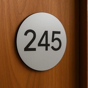 Könnte beinhalten: Rundes, silberfarbenes Türschild mit der Zahl 245 in fetter schwarzer Schrift. Das Schild ist an einer Holztür befestigt und hat einen schwarzen Rand.