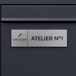 Könnte beinhalten: Ein rechteckiges silbernes Schild mit dem Text "ATELIER N°1" in Schwarz und dem "UNISIGN"-Logo mit einem Stiftsymbol. Das Schild ist auf einer dunkelgrauen Oberfläche montiert.