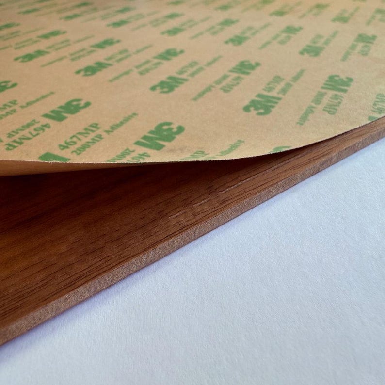 May include: Close-up of a brown, wood-grain textured material with a partially peeled adhesive backing. The backing is tan with green text and logos. The adhesive is labeled "467MP 200MP Adhesive".