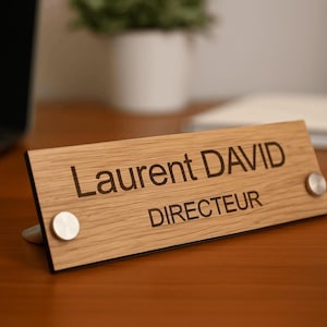 May include: Wooden desk nameplate with the name "Laurent David" and the title "Directeur" engraved in black. The nameplate is rectangular, with silver-coloured circular fasteners.