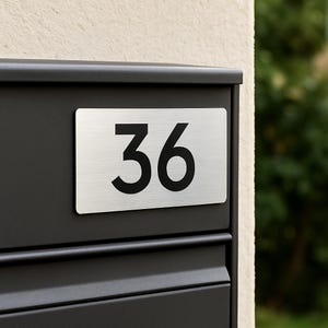 May include: A rectangular address plaque displaying the number 36 in black on a brushed metal surface. The plaque is affixed to a black mailbox. The background features a beige wall and green foliage.