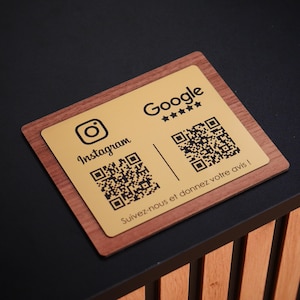 Peut inclure: Une carte de visite dorée avec le logo Instagram et un code QR à gauche, et le logo Google avec des étoiles et un code QR à droite. La carte est posée sur une base en bois.