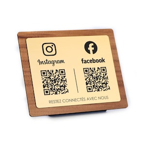 Puede incluir: Un letrero de madera con una placa metálica dorada. La placa presenta los logotipos de Instagram y Facebook, códigos QR y el texto "Restez connectés avec nous". El letrero es de forma cuadrada.