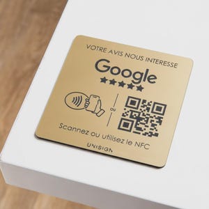 Peut inclure: Un panneau carré doré avec du texte et des graphiques noirs. Le panneau présente le logo Google, cinq étoiles, un code QR et le texte "Scannez ou utilisez le NFC". Le panneau est sur une surface blanche.
