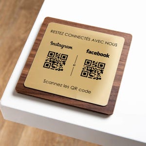 Peut inclure: Un panneau en bois et doré avec le texte "RESTEZ CONNECTÉS AVEC NOUS". Il présente les logos Instagram et Facebook avec des codes QR, et le texte "Scannez les QR code". Le panneau est posé sur une surface blanche.