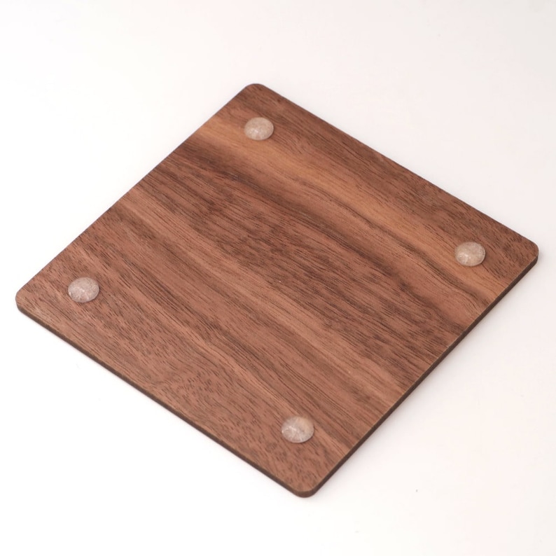May include: A square, wooden coaster with a rich brown color and visible wood grain. The coaster has four small, clear, rounded feet on the corners. The coaster is designed to protect surfaces from drink condensation.