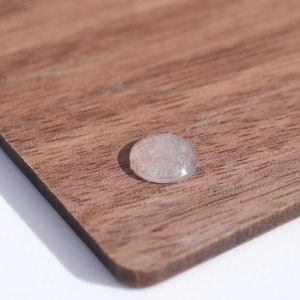 May include: A small, clear, rounded object with a slightly pinkish hue rests on a wooden surface. The wood has a rich, brown color with visible grain patterns. The object appears to be smooth and translucent, reflecting light.