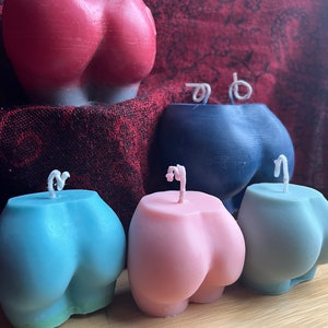 May include: Four butt-shaped candles in different colors: red, blue, light blue, and pink. The candles are arranged in a row on a wooden surface.