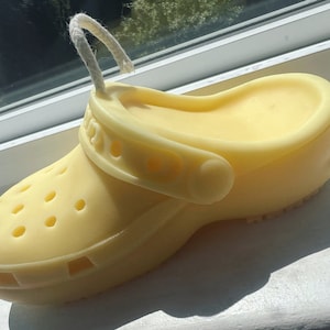 Puede incluir: Un jabón amarillo claro con forma de zapato Crocs, con los agujeros y la correa característicos. Una cuerda blanca está unida a la parte superior. El jabón está sobre una superficie blanca.