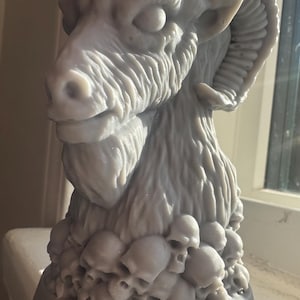 May include: A gray sculpture of a ram's head with large curved horns. The ram's head is detailed with textured fur and a beard. The base of the sculpture is adorned with a cluster of small skulls. The sculpture is displayed on a windowsill.