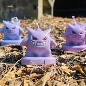 Puede incluir: Tres figuras de velas moradas del personaje Pokémon Gengar. Cada Gengar tiene una sonrisa blanca y ojos rojos. Las velas están sentadas en una cama de astillas de madera marrón.