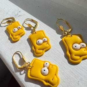May include: A set of four yellow cartoon character earrings with gold hooks. Each earring features a yellow face with white eyes and a yellow mouth.