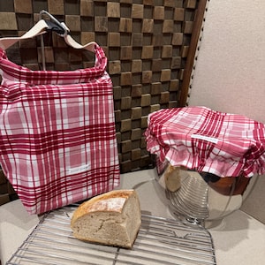 May include: A red and white plaid fabric bread bag with a black buckle closure and a matching red and white plaid fabric bowl cover with an elastic edge. Both items feature a small white label with the text sweet slice. A loaf of bread sits nearby.