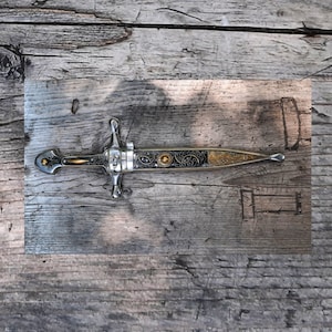 May include: A silver dagger with a black and gold handle and intricate details. The dagger is lying on a wooden surface.